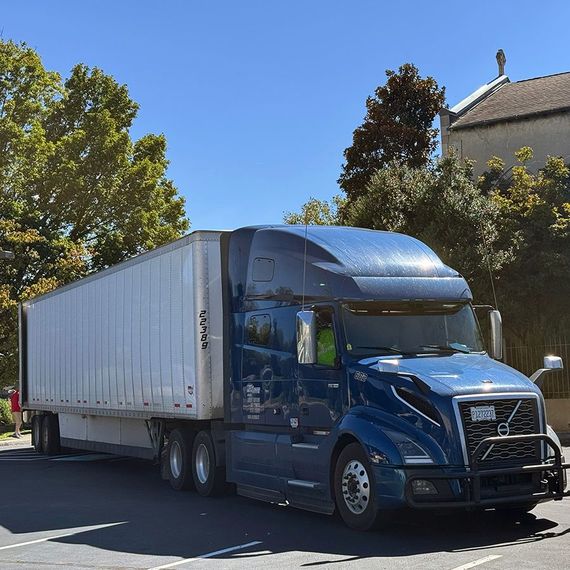 Blue semi-truck with white trailer parked in front of trees and a building on a sunny day.