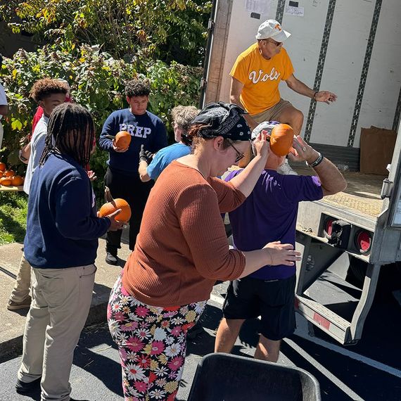 People unloading pumpkins from a truck. Outdoors, sunny. Volunteers in casual attire pass pumpkins to each other.
