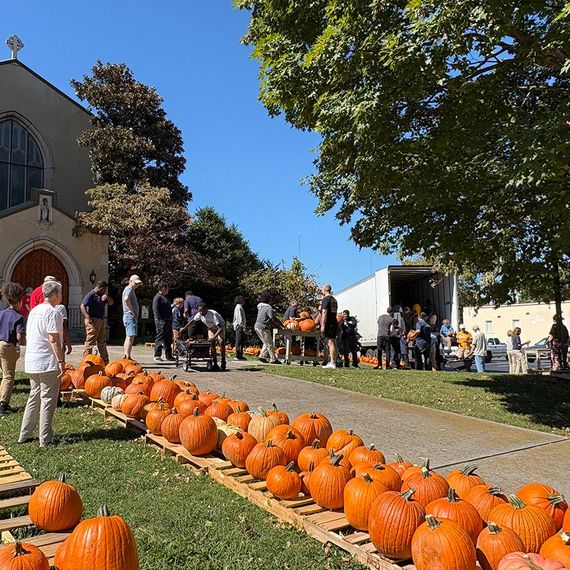 People at a church pumpkin patch: pumpkins on pallets, unloading truck, sunny day.