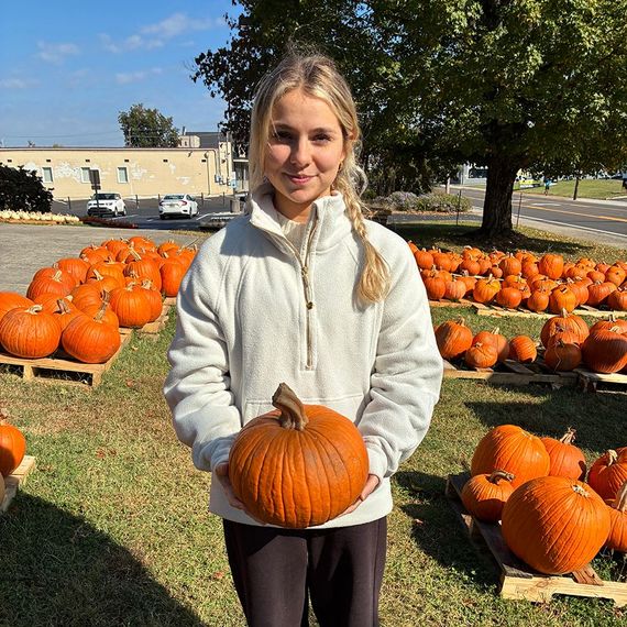 Blonde person in white fleece holds pumpkin in a pumpkin patch. Pallets of pumpkins in the background. Sunny day.