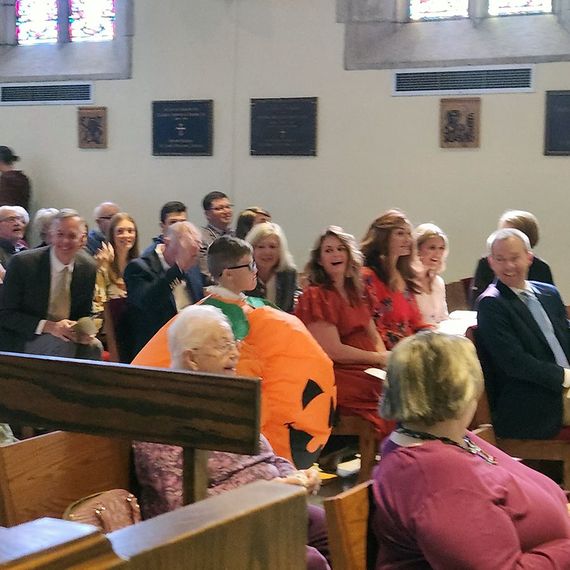 People seated in a church; a person in a pumpkin costume among them.