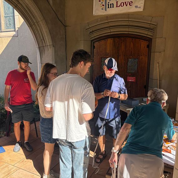 Group of people outdoors near a wooden door and archway. Some are looking at a table with items.