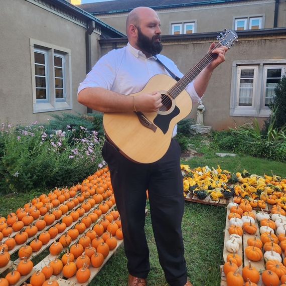Man playing acoustic guitar outdoors amidst rows of pumpkins.