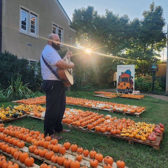 Man with guitar plays surrounded by pumpkins in a garden, with sunlight beaming through the trees.