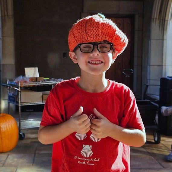 Boy wearing pumpkin hat and red shirt, giving thumbs up, smiling. Indoors with pumpkins.