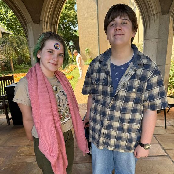 Two young people stand outside; one with green hair, a pink scarf, and a sticker; the other in a plaid shirt and watch.