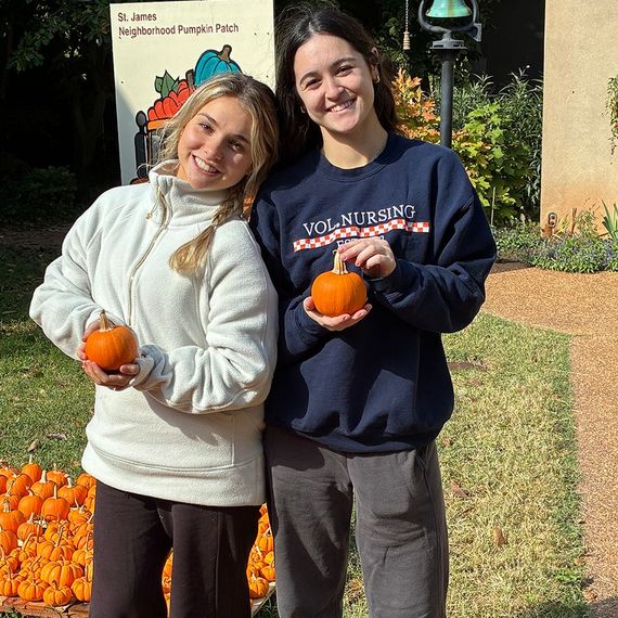 Two smiling people at a pumpkin patch, each holding a small pumpkin. They're outdoors in front of pumpkins and a sign.