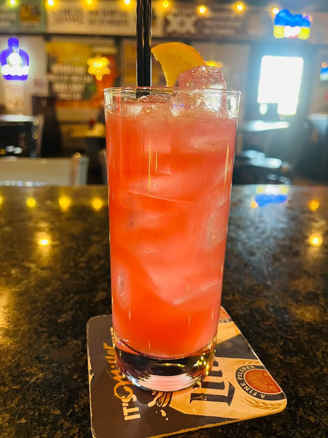 A tall glass of red liquid with ice and a straw on a coaster on a bar.