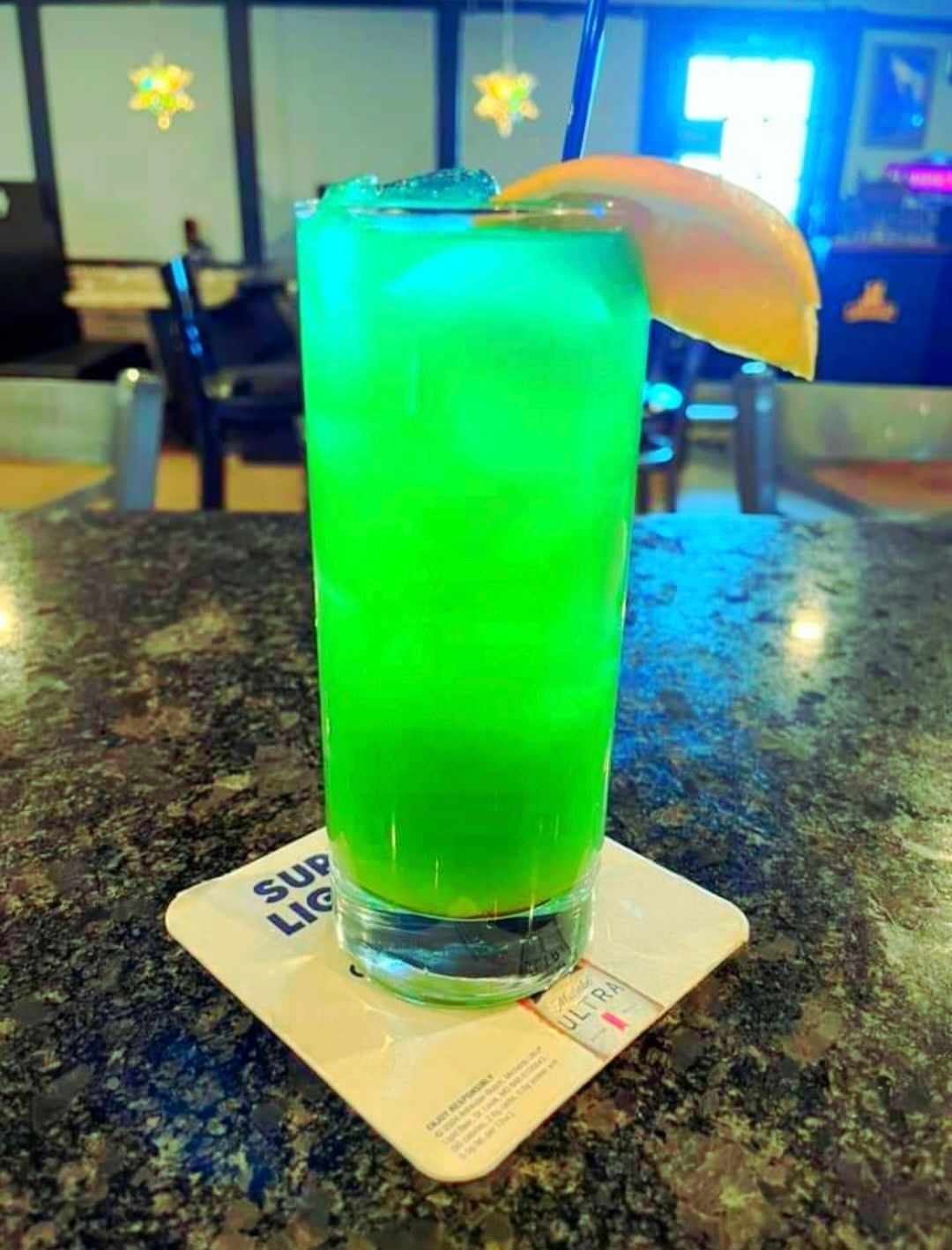 A green drink with a slice of orange on a coaster on a table.