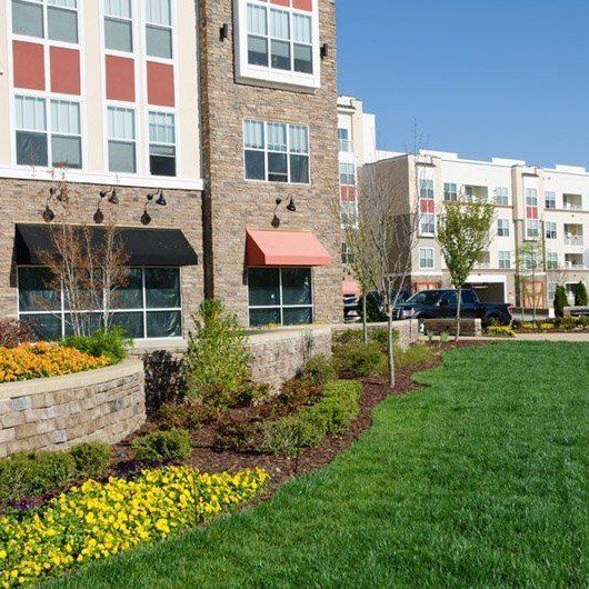 Apartment complex exterior with stone facade, awnings, landscaping, and a grassy lawn.