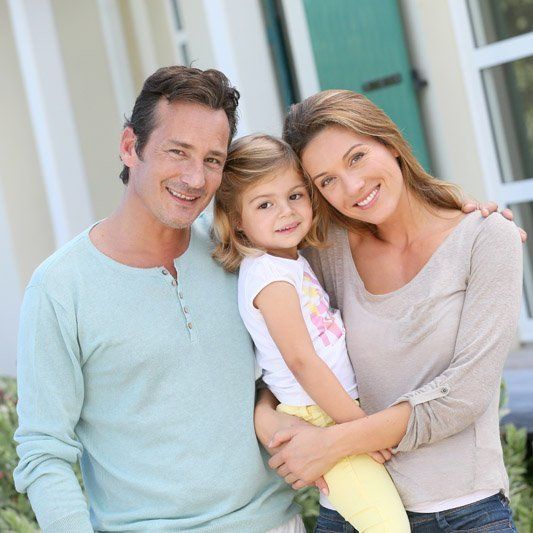 Family of three smiling, posing in front of a house.