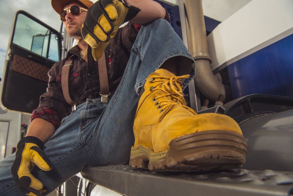 A man wearing a pair of yellow work boots is sitting on the steps of a semi truck.