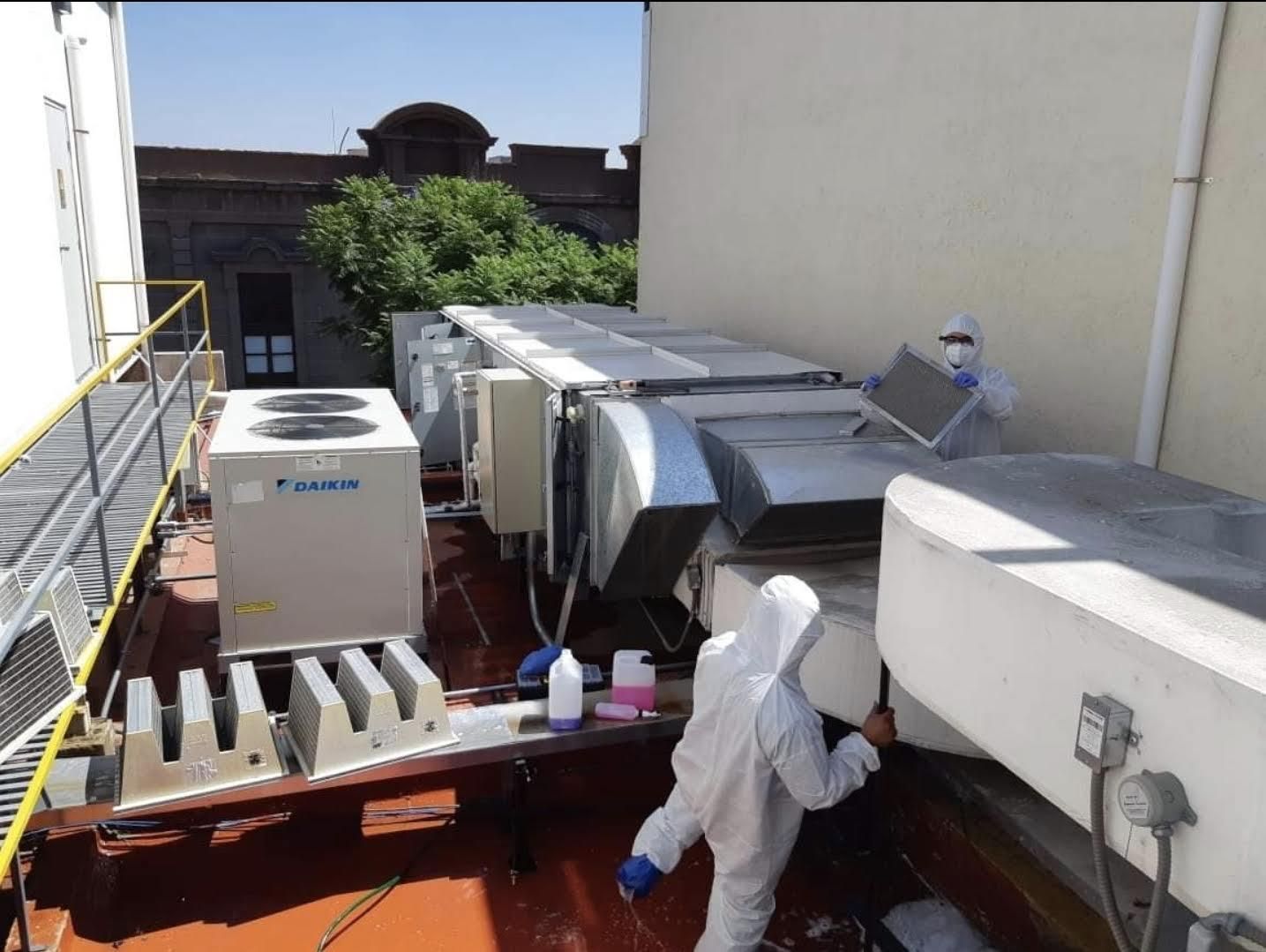 Dos técnicos con trajes protectores blancos limpian y dan mantenimiento a unidades industriales de climatización en la azotea de un edificio.