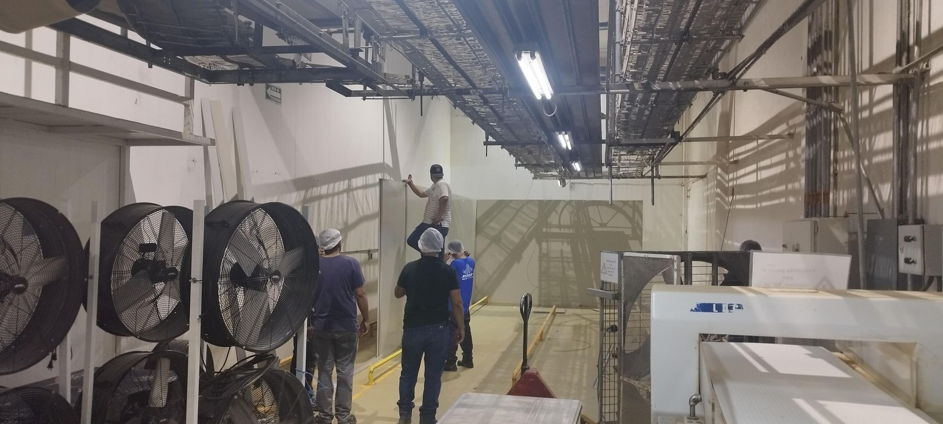 En una instalación industrial, los trabajadores instalan paneles de pared rodeados de grandes ventiladores y conductos eléctricos montados en el techo.