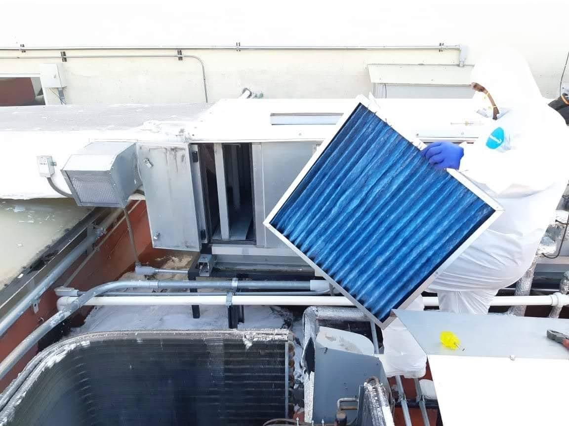 Una persona con un traje protector reemplaza un filtro de aire cuadrado azul en una unidad de climatización en una azotea.