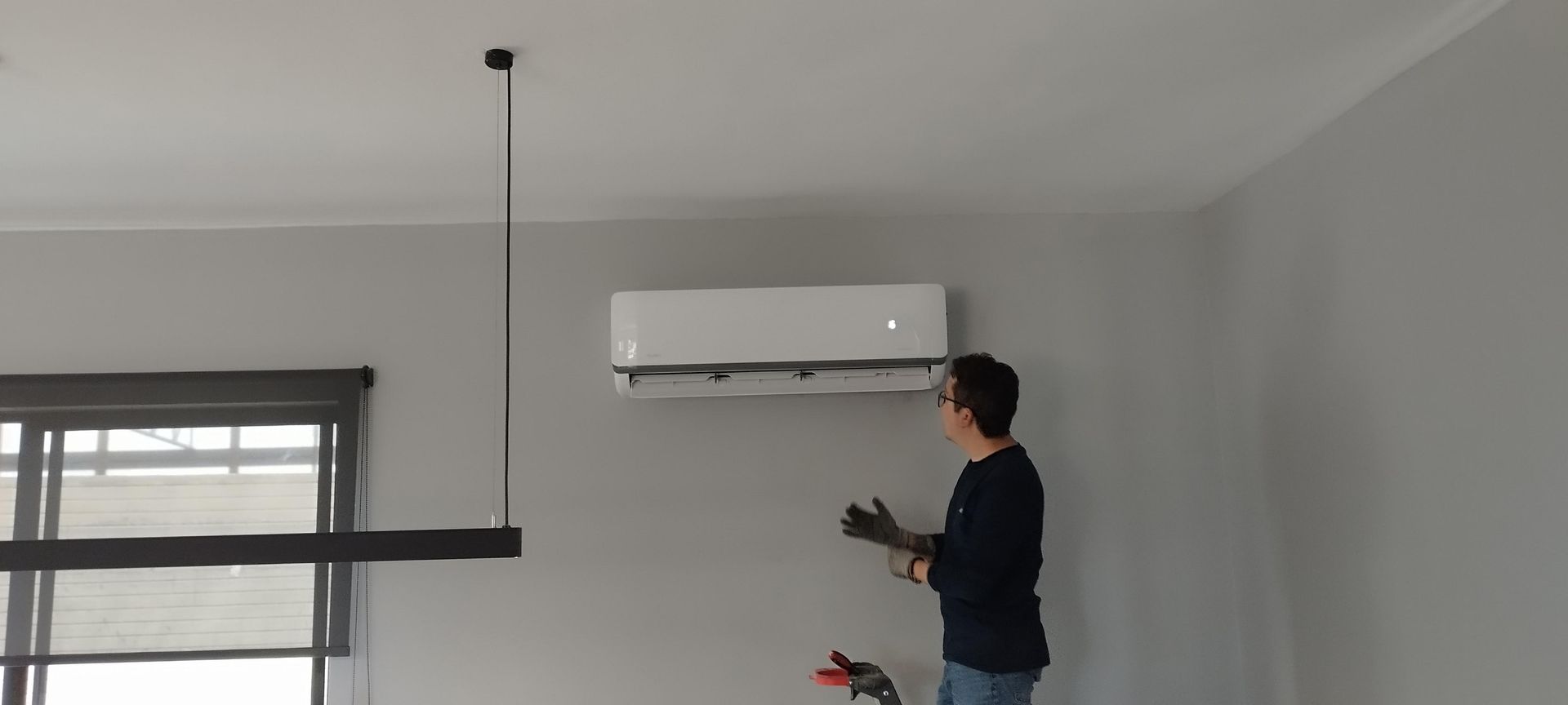 Una persona con una camisa oscura de manga larga y guantes instala una unidad de aire acondicionado blanca montada en la pared de una habitación.