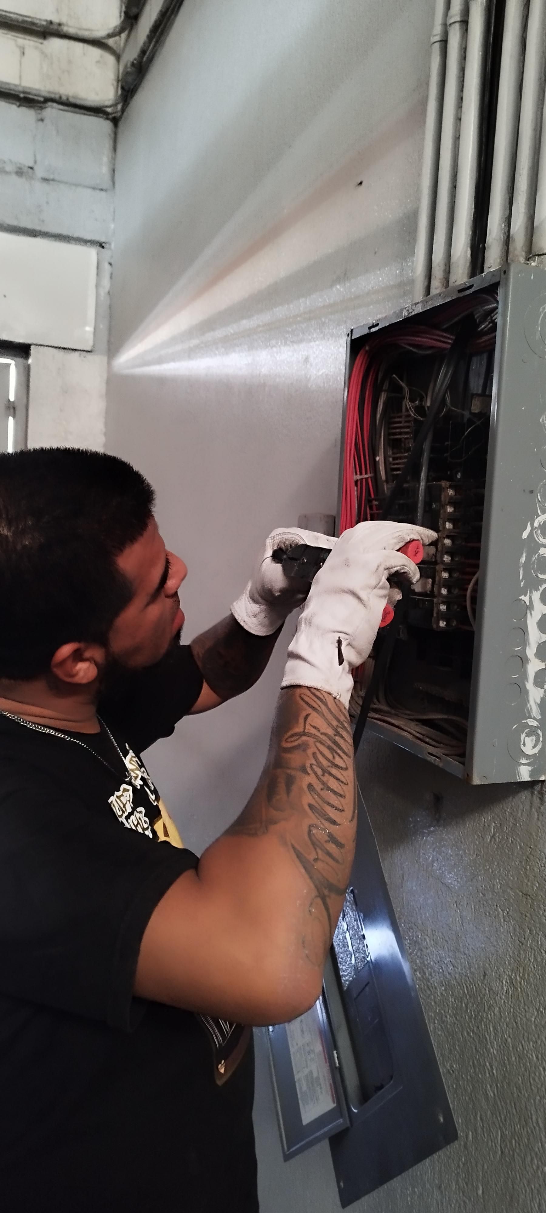 Un técnico con tatuajes en el brazo, que lleva guantes blancos, trabaja en el panel abierto de una caja de disyuntores eléctricos.