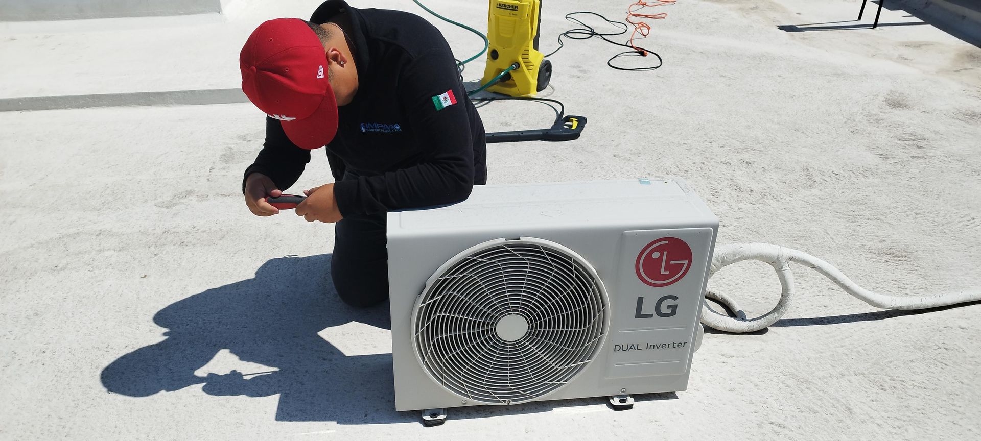 Una persona con una gorra roja arrodillada en una azotea mientras trabaja en una unidad de aire acondicionado LG.