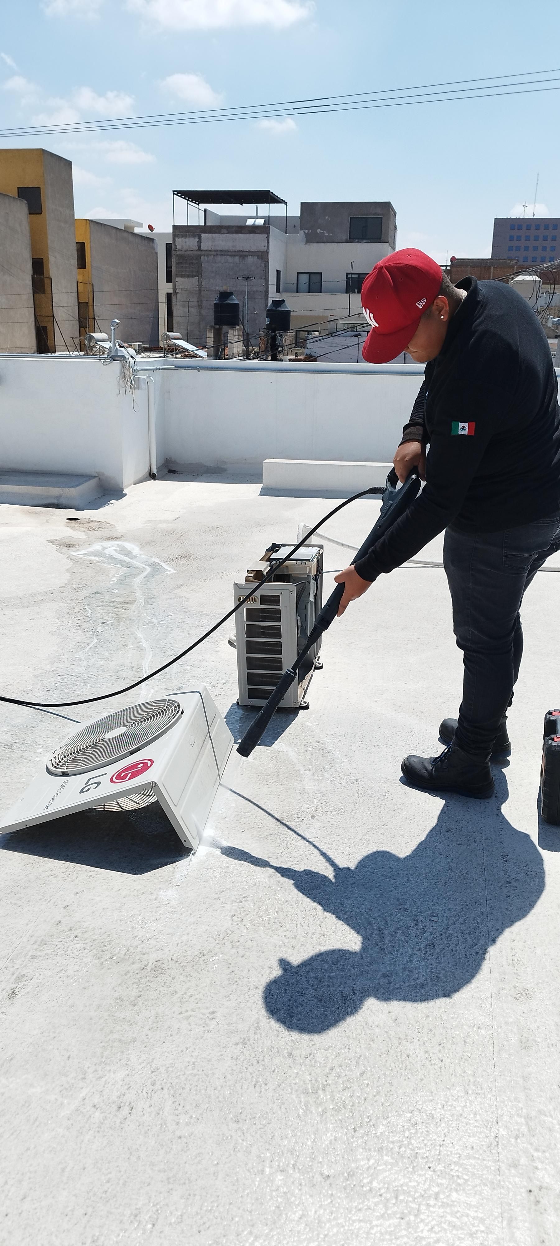 Un técnico con gorro rojo utiliza una hidrolimpiadora de alta presión para limpiar una unidad de aire acondicionado en una azotea.