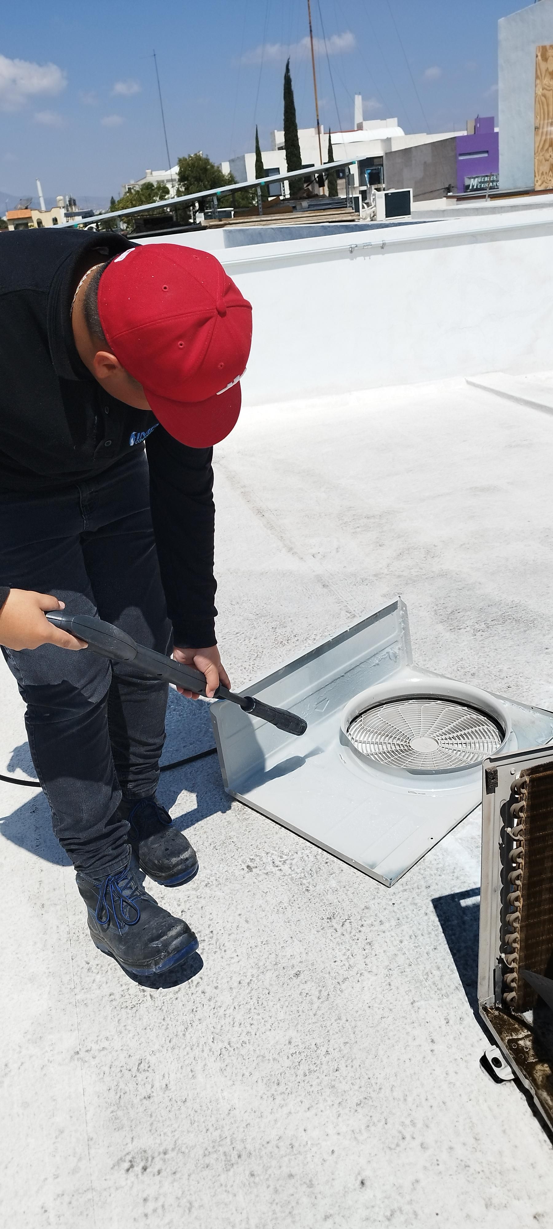 Un técnico con gorro rojo y uniforme oscuro utiliza una herramienta en una rejilla de ventilación del techo, cerca de una unidad de climatización, en un tejado plano blanco.