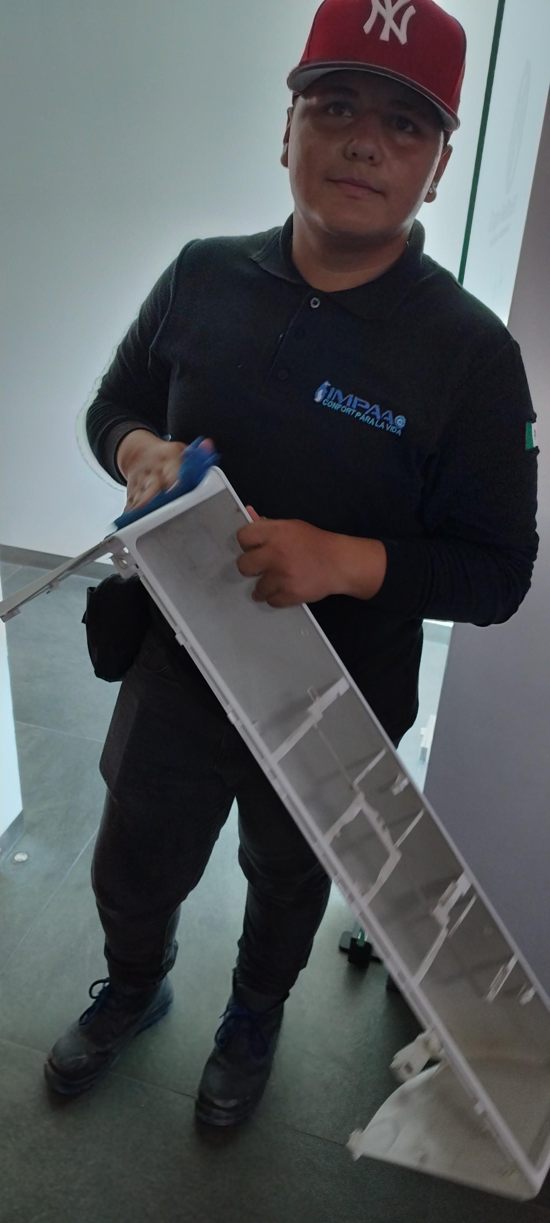 Una persona con gorra roja y uniforme oscuro sostiene un filtro de aire acondicionado largo y rectangular para limpiarlo.