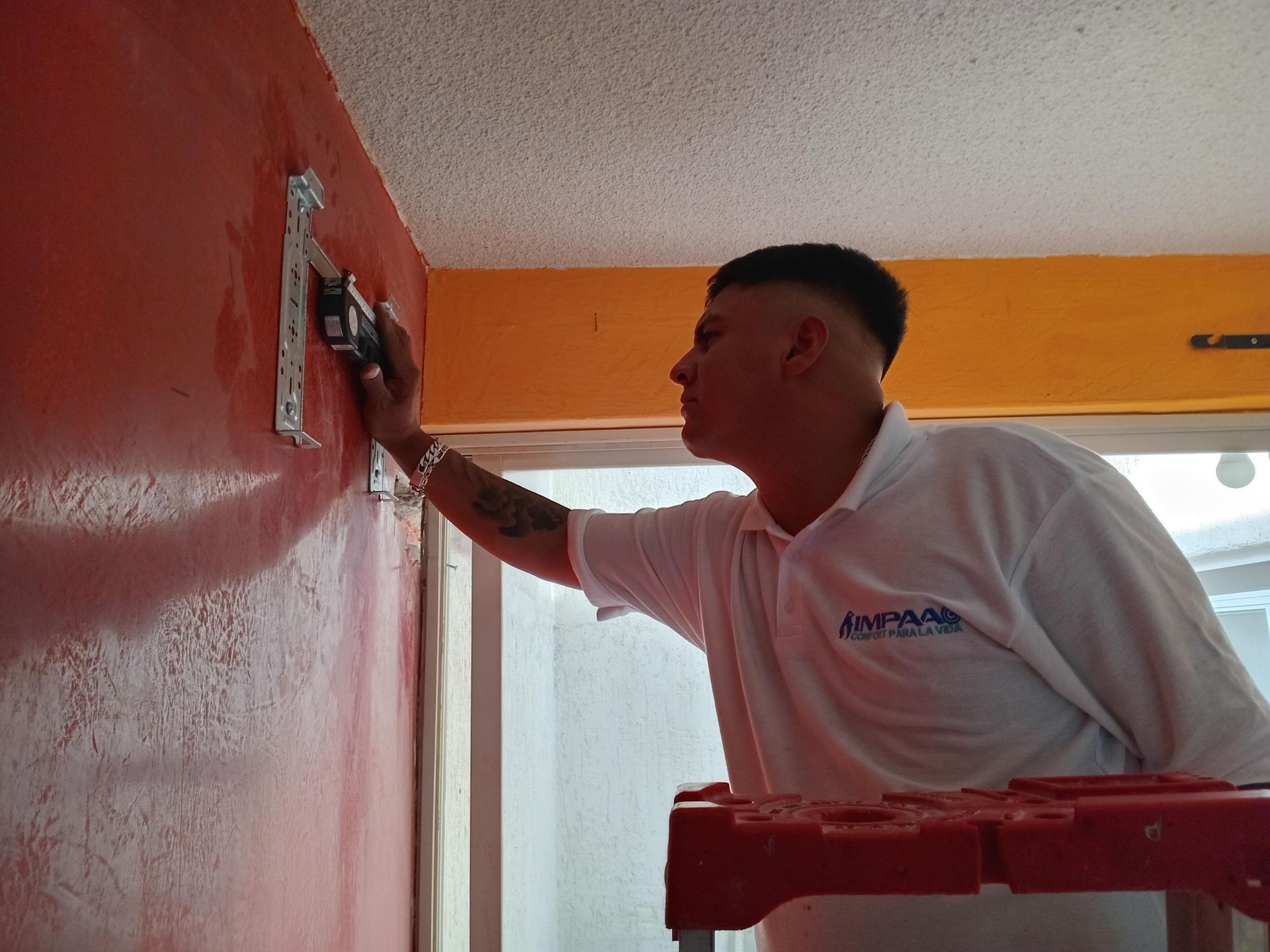 Una persona con una camisa polo blanca utiliza un nivel para instalar un soporte de montaje metálico en una pared rojiza.