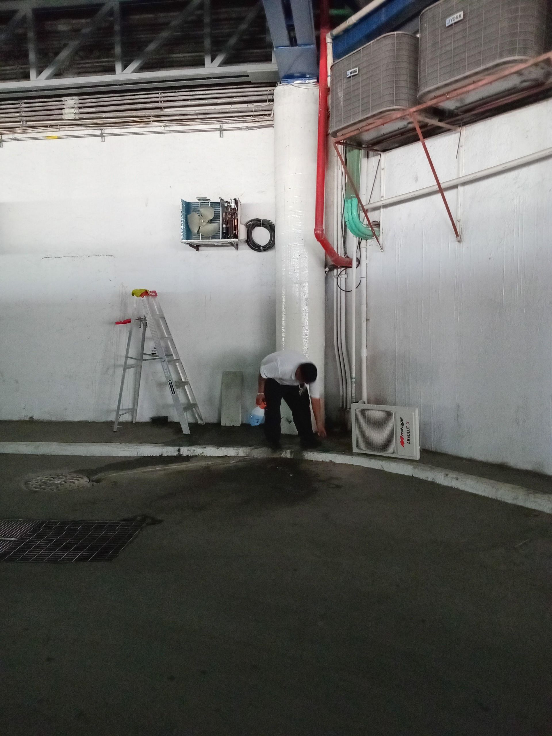 Una persona vestida con ropa oscura trabaja cerca de un gran pilar blanco en una habitación con paredes blancas, una escalera metálica y tuberías de servicios públicos.
