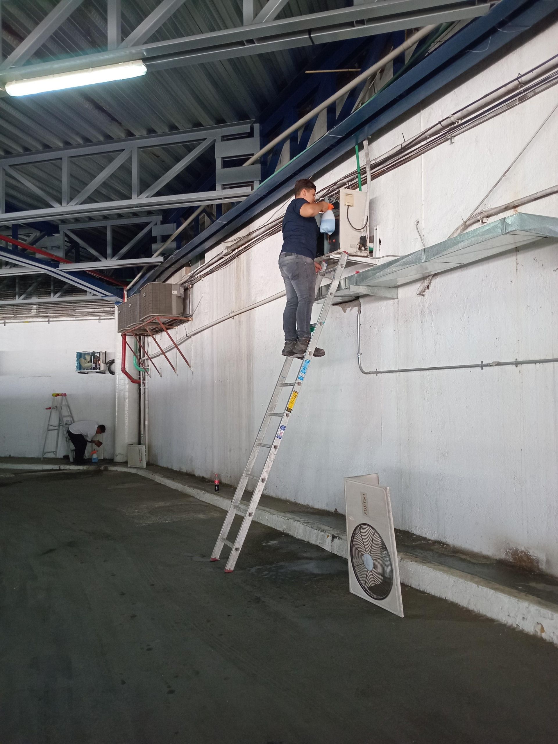 Un trabajador se encuentra subido a una escalera alta, reparando equipos eléctricos montados en una pared blanca de un gran edificio industrial.