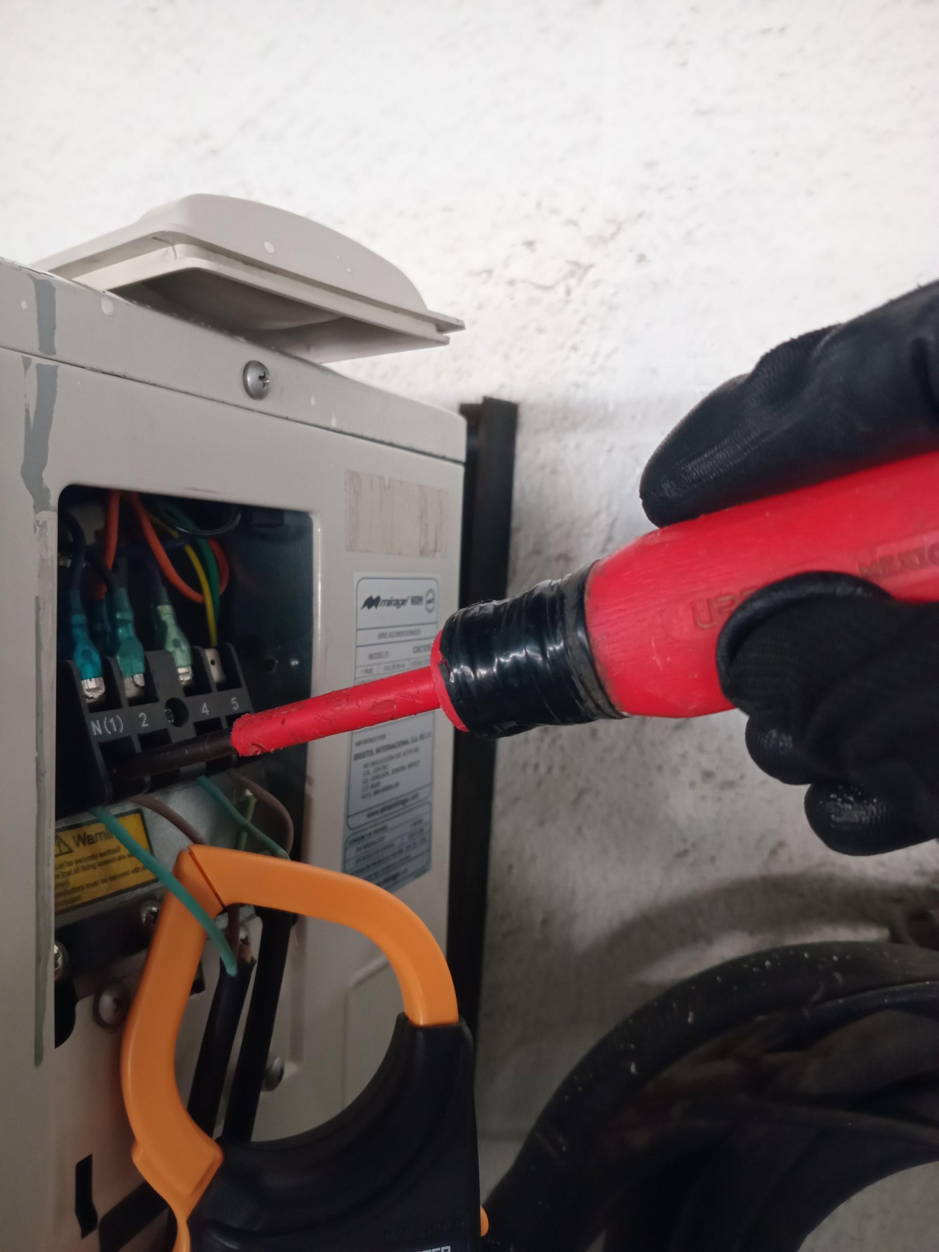 Una mano enguantada utiliza un comprobador de voltaje en el cableado de una unidad de aire acondicionado exterior mientras se le conecta un amperímetro de pinza.