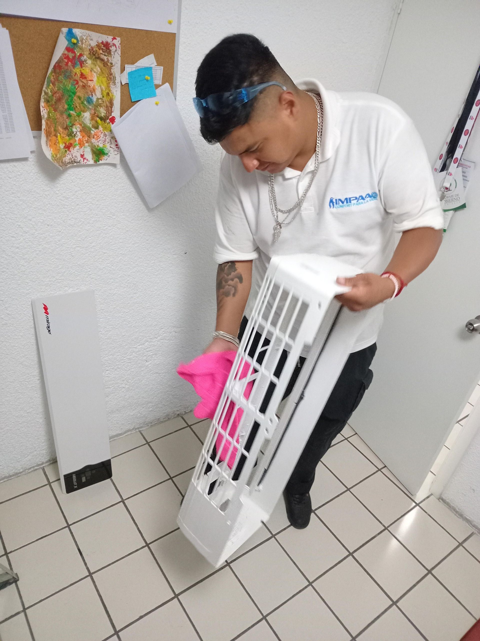Una persona con uniforme blanco utiliza un paño rosa para limpiar el panel frontal de plástico blanco de una unidad de aire acondicionado interior.