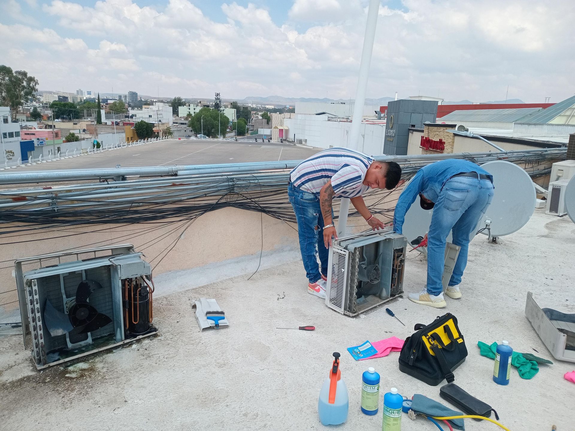 Dos personas vestidas con ropa informal trabajan en unidades de aire acondicionado desmontadas en una azotea con vistas a la ciudad.