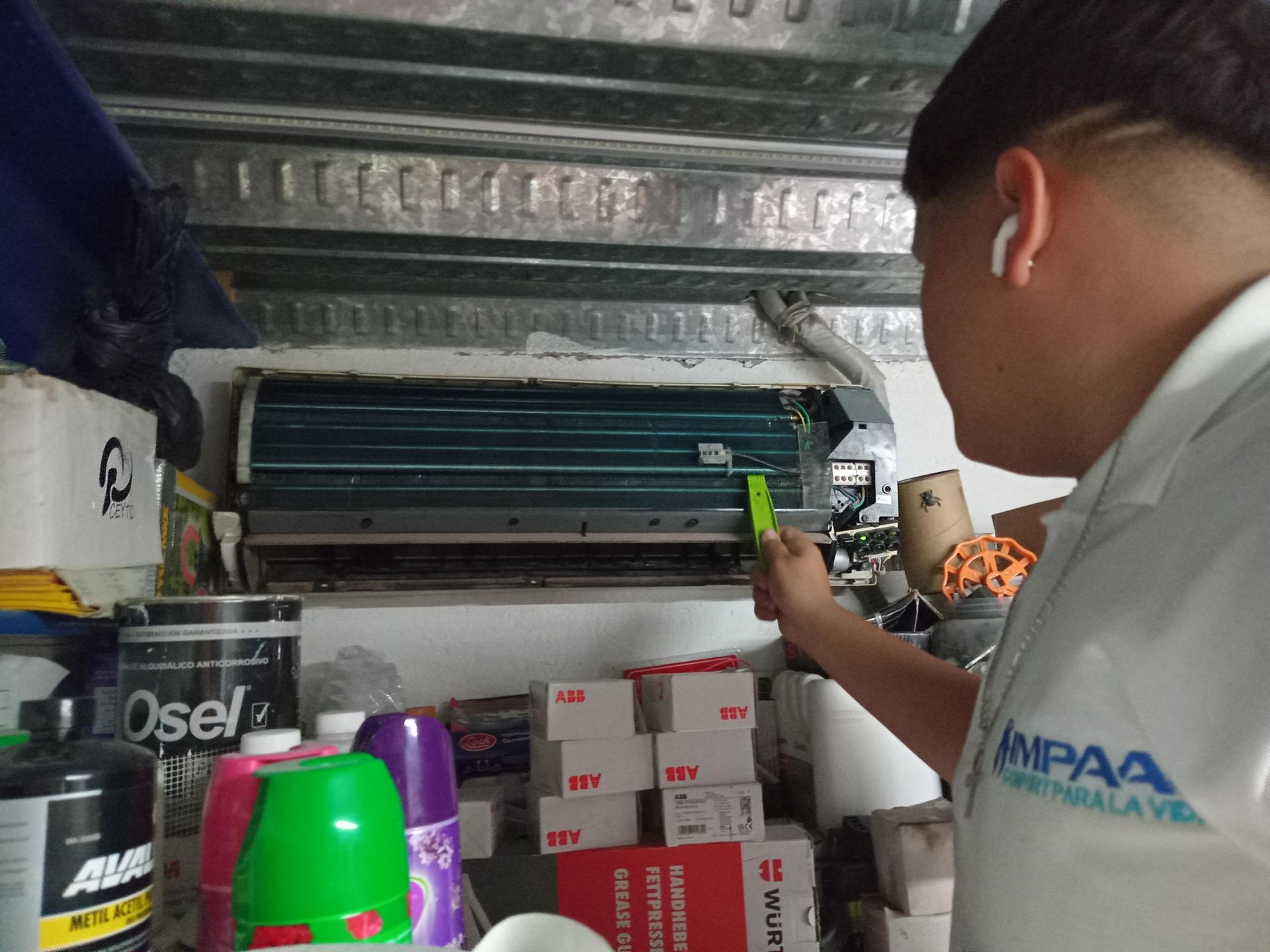 Un técnico con camisa blanca trabaja en una unidad de aire acondicionado montada en la pared en una habitación desordenada.