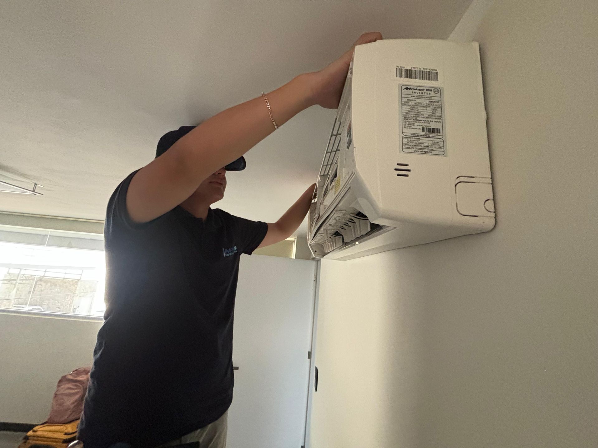 Una persona con una camiseta negra está subida a una escalera, instalando o reparando una unidad de aire acondicionado blanca montada en la pared.