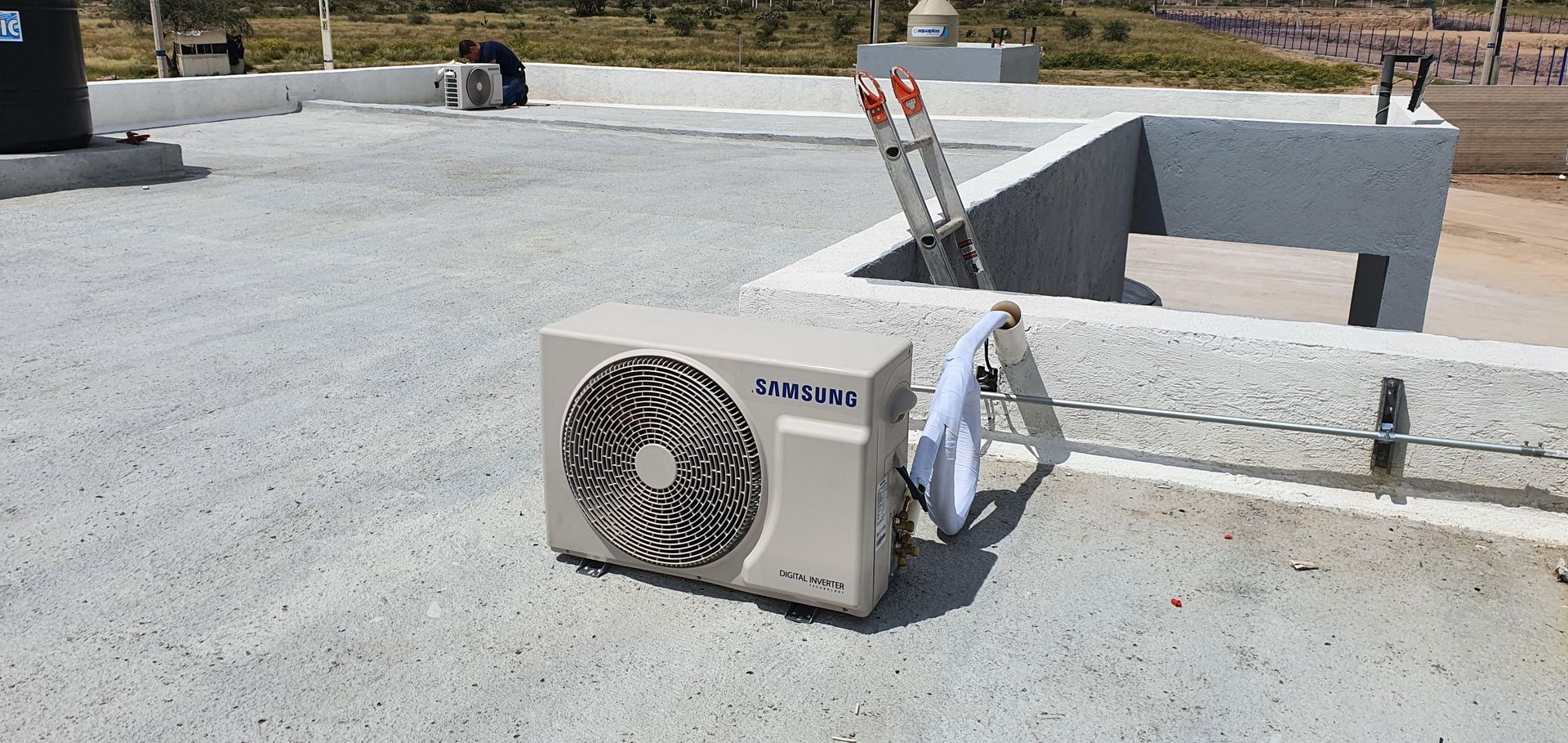 Una unidad de aire acondicionado exterior de color claro se encuentra sobre una azotea plana de hormigón gris, bajo la brillante luz del sol.