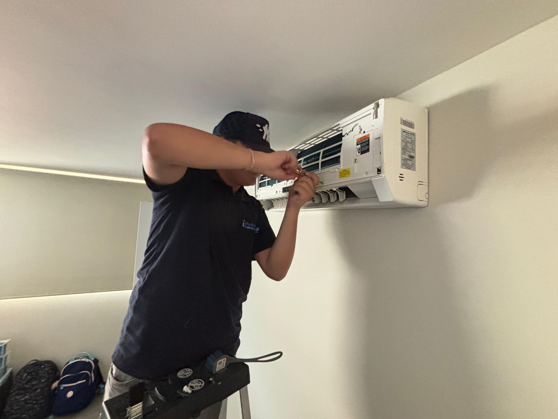 Un técnico con camisa y gorra negras utiliza un destornillador para dar mantenimiento a una unidad de aire acondicionado blanca montada en la pared.