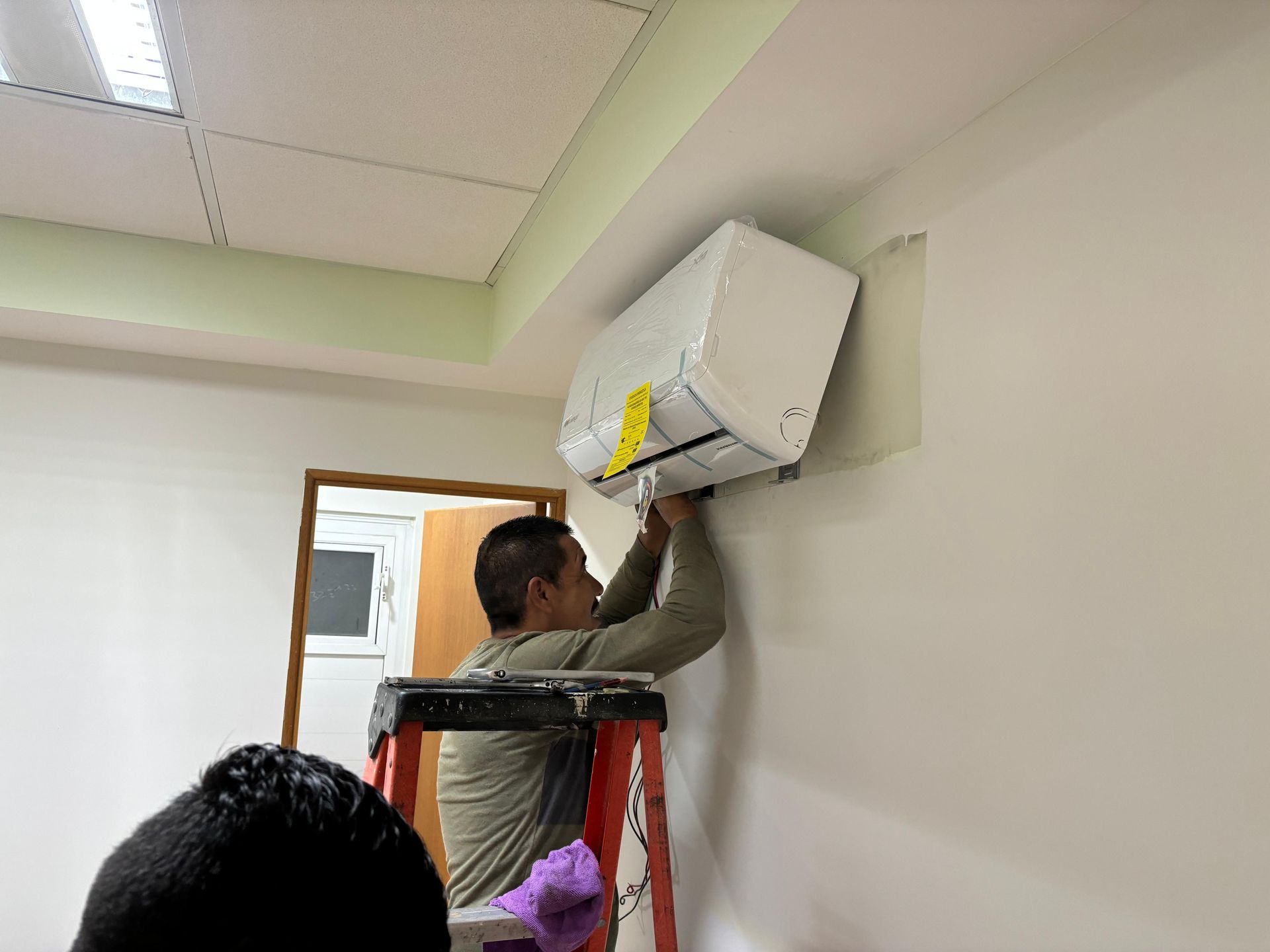Un técnico se encuentra subido a una escalera, instalando o reparando una unidad de aire acondicionado blanca montada en la pared de una oficina.