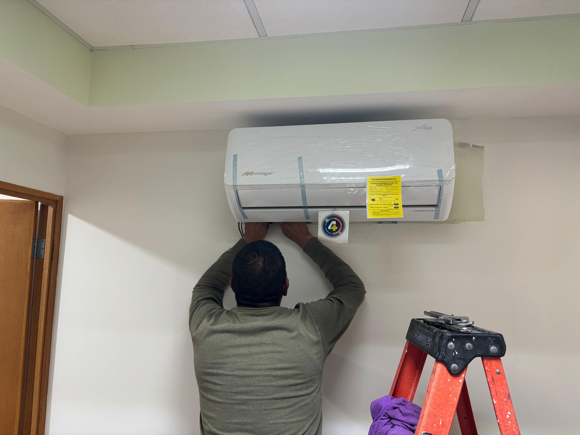 Una persona, subida a una escalera, instala una unidad de aire acondicionado blanca de pared en una habitación interior.