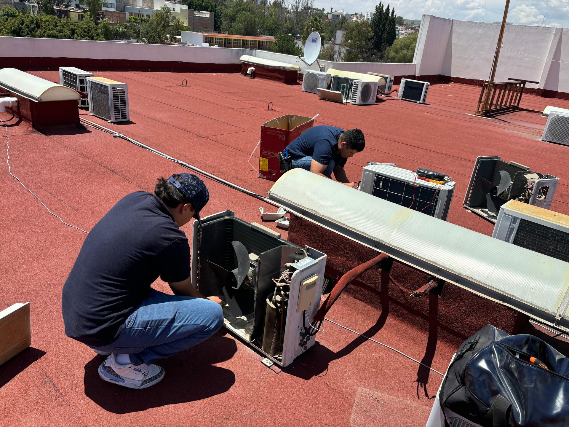 Dos técnicos reparan unidades de aire acondicionado en una azotea con superficie roja durante el día.