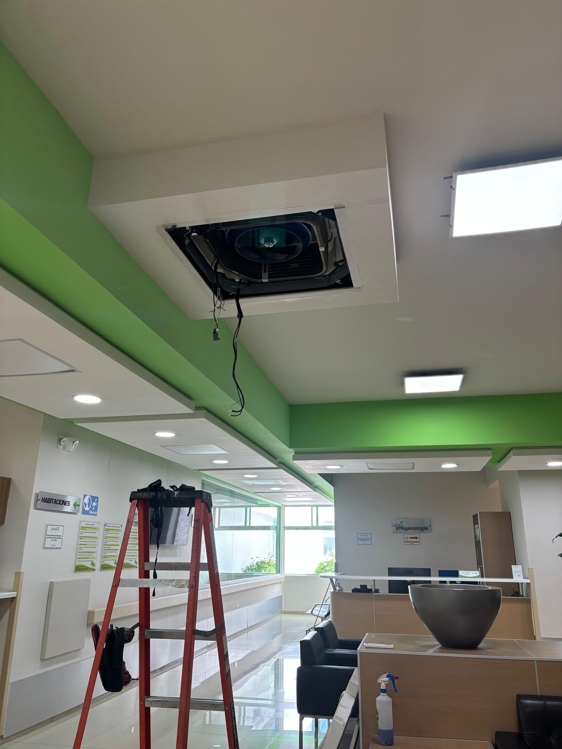 Una escalera roja colocada debajo de una unidad de aire acondicionado montada en el techo, a la que se le ha retirado la cubierta, en un espacio de oficina.