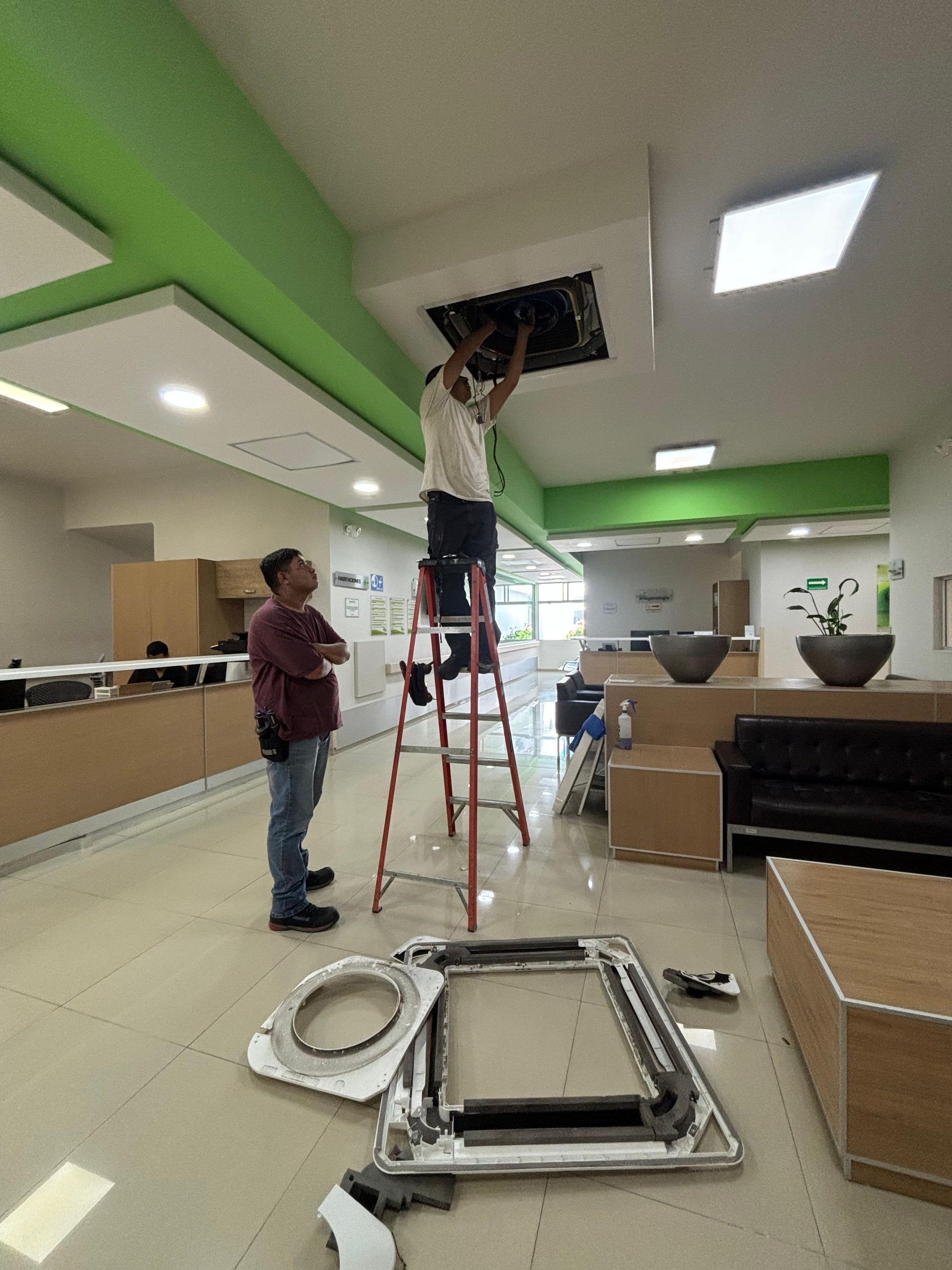 Dos trabajadores reparan una unidad de climatización instalada en el techo del vestíbulo de una oficina; uno está subido a una escalera mientras el otro observa.