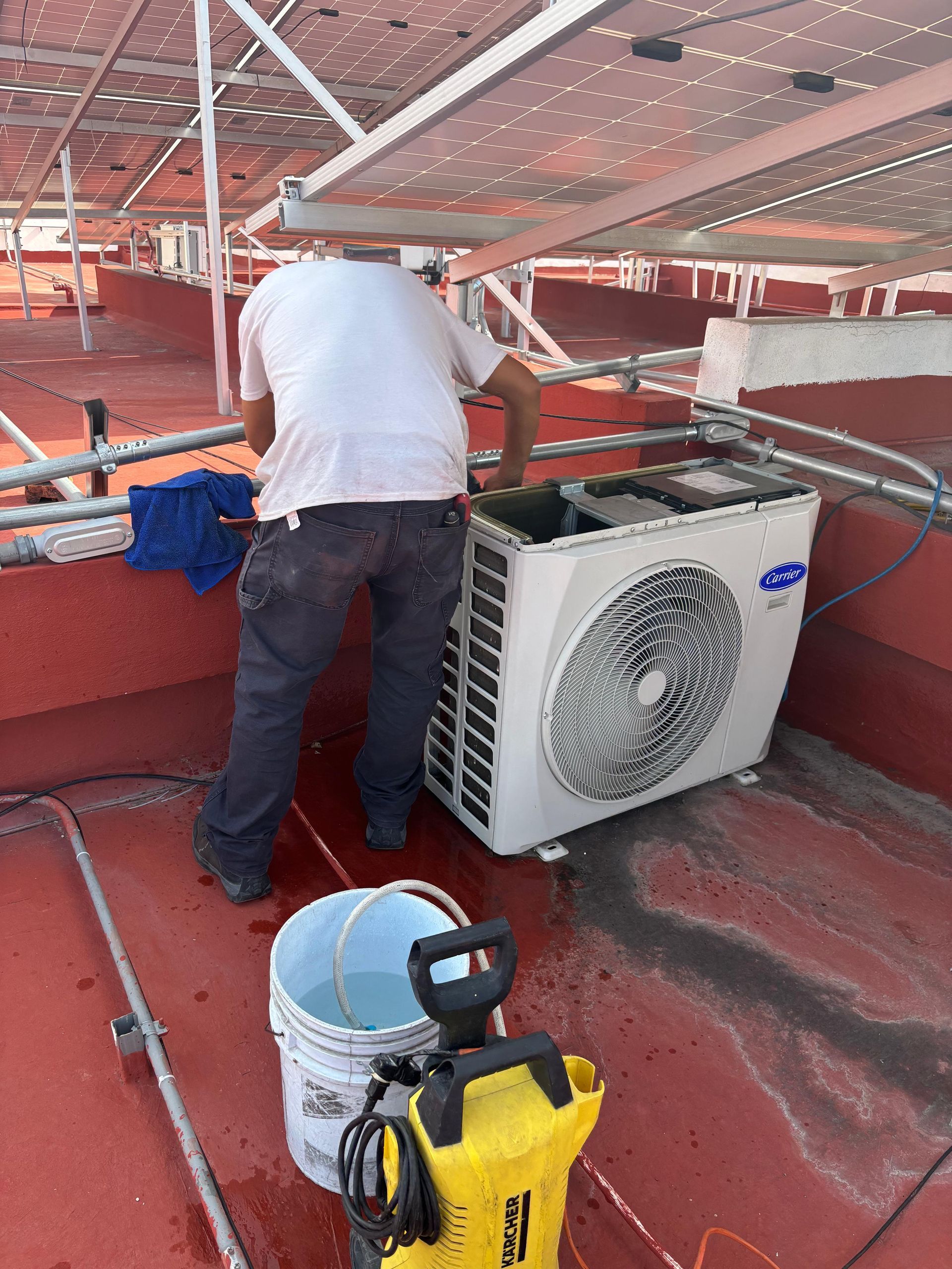 Un técnico limpia una unidad de aire acondicionado exterior en un tejado rojo, debajo de paneles solares, utilizando una hidrolimpiadora amarilla.