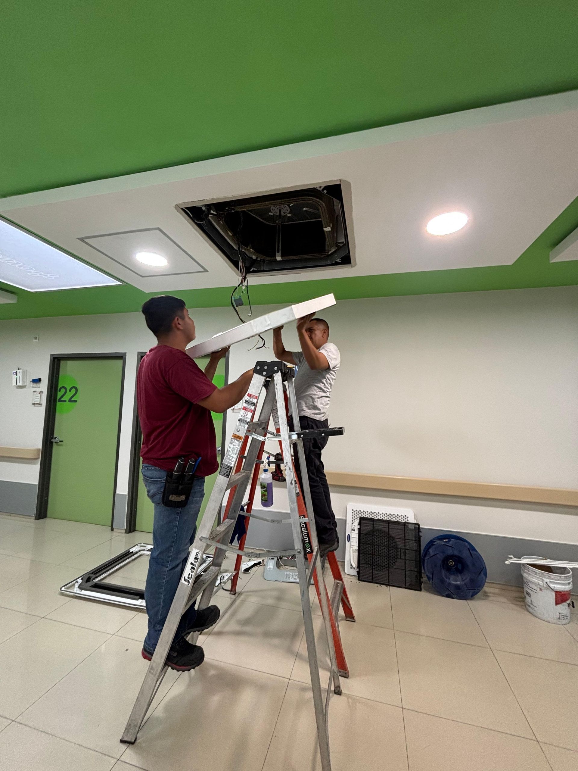 Dos trabajadores subidos a una escalera instalan una lámpara de techo cuadrada en una oficina interior luminosa y moderna.