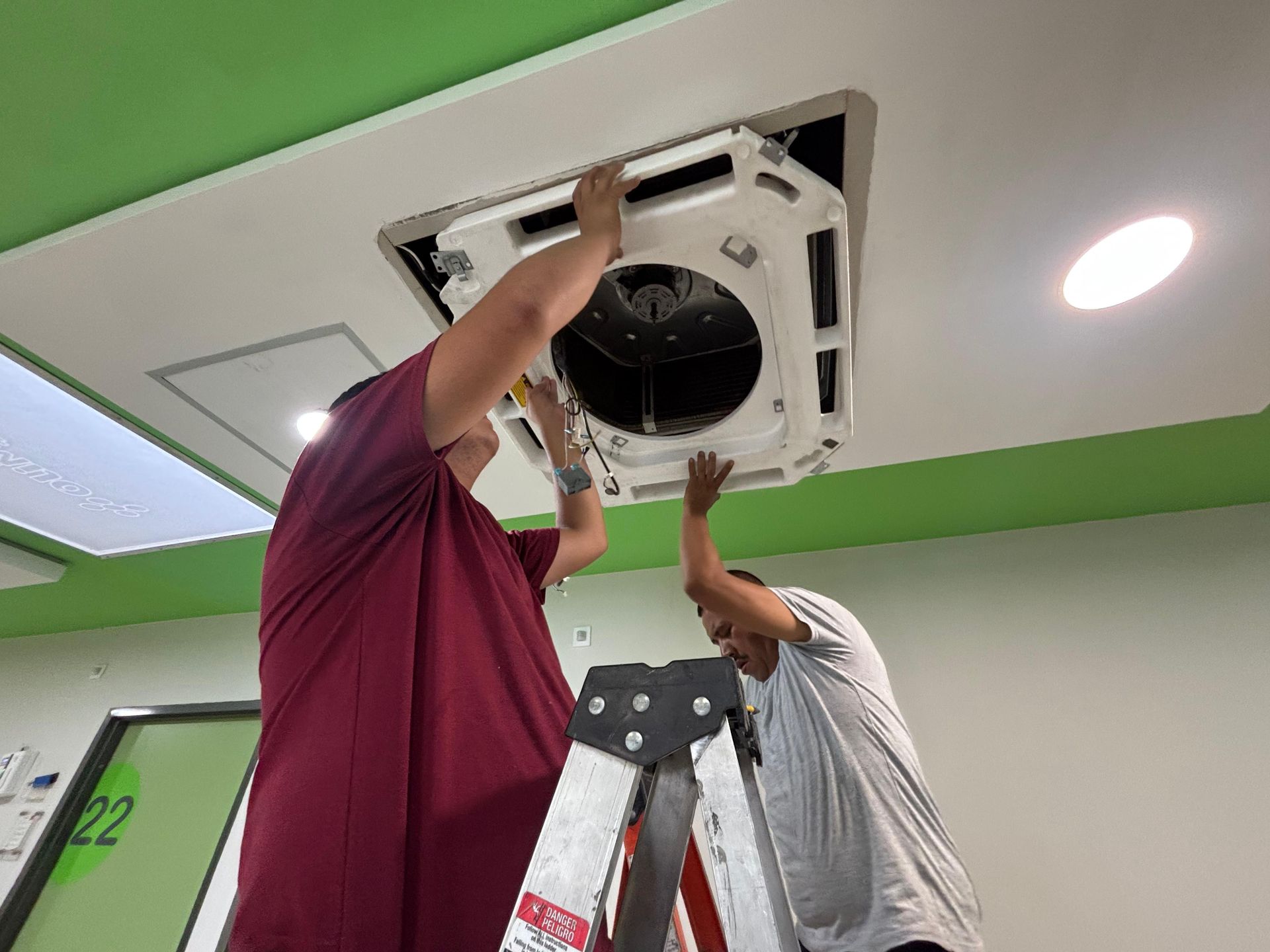 Dos trabajadores subidos a una escalera instalando o reparando una unidad de climatización montada en el techo en un entorno similar a una oficina.