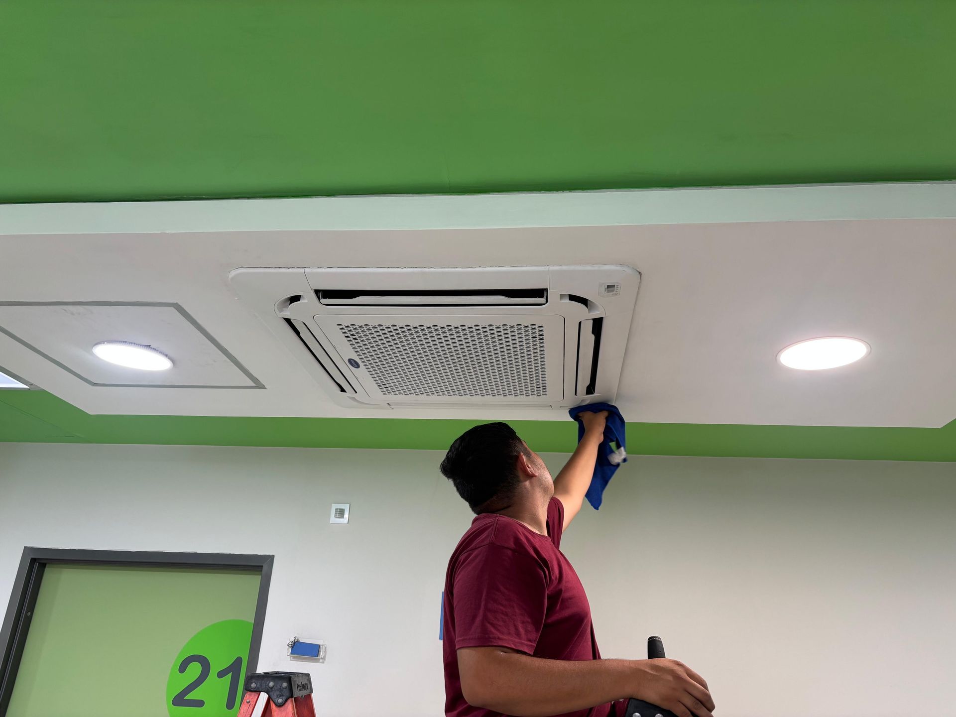 Una persona con una camisa granate está subida a una escalera, limpiando una unidad de aire acondicionado montada en el techo con un paño azul.