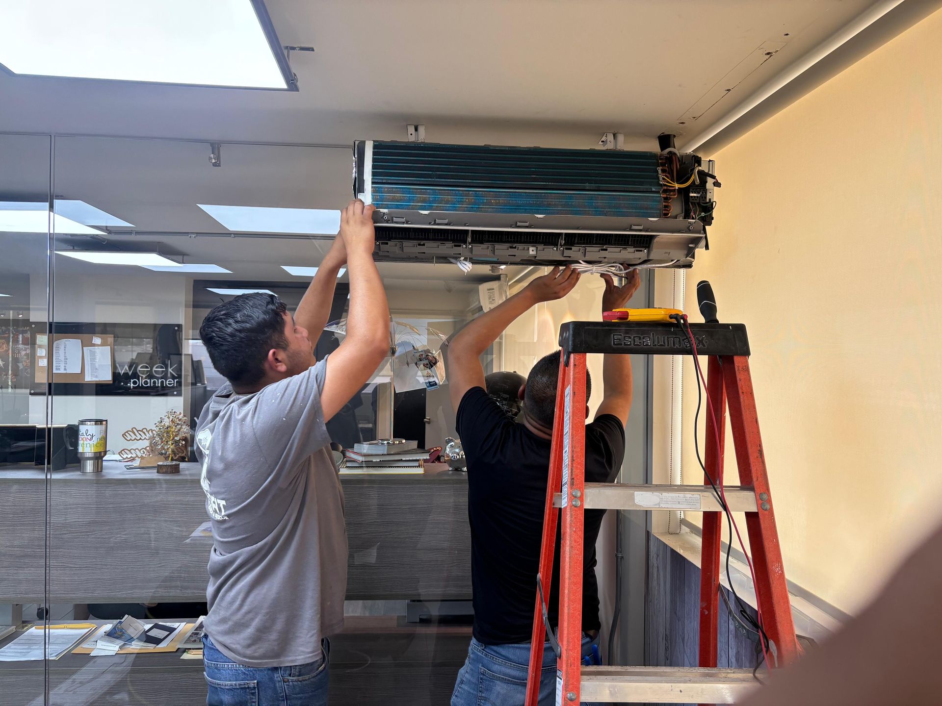 Dos técnicos están subidos a una escalera y al suelo, trabajando en una unidad de aire acondicionado abierta montada en el techo.