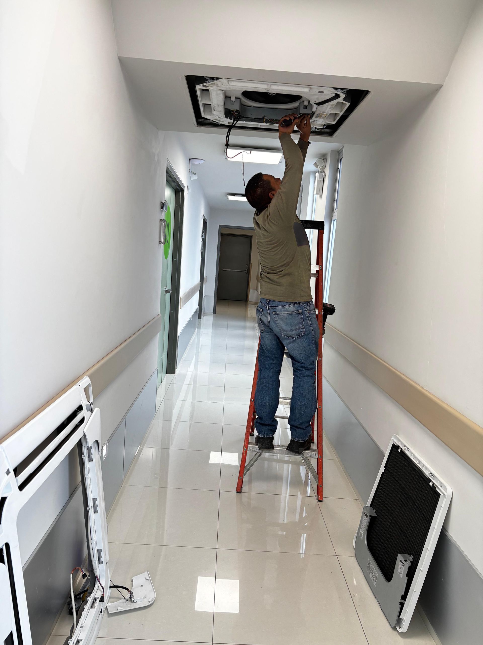 Una persona se encuentra subida a una escalera en un pasillo blanco, reparando una unidad de aire acondicionado instalada en el techo.