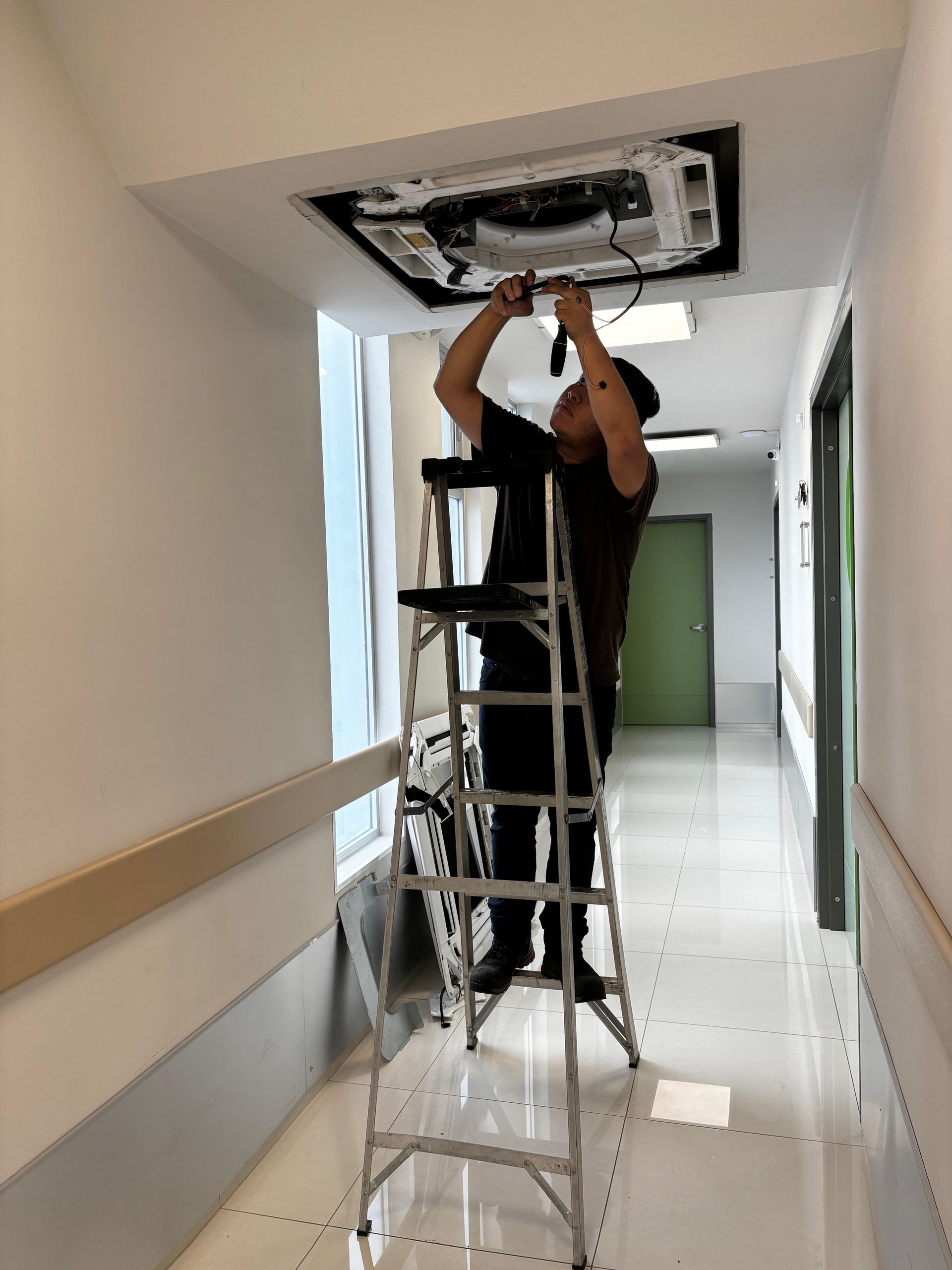 Una persona de pie sobre una escalera en un pasillo, reparando o dando mantenimiento a una unidad de aire acondicionado montada en el techo.