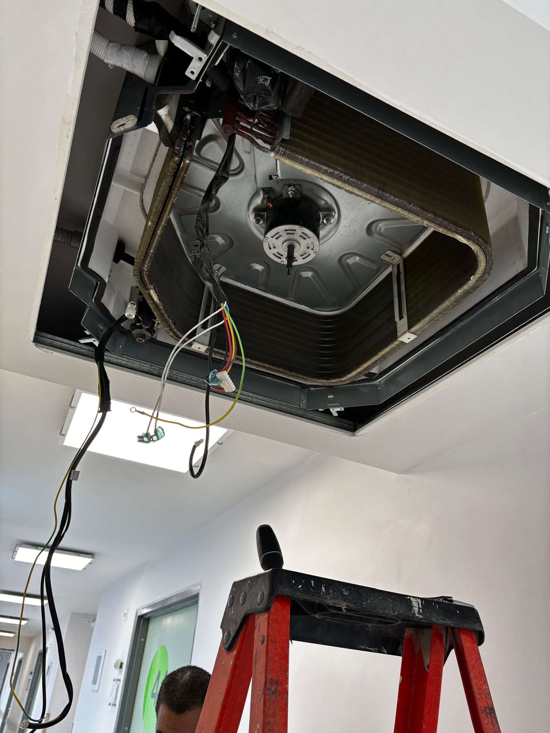 Una unidad de aire acondicionado abierta montada en el techo, en proceso de mantenimiento, con una escalera roja debajo.