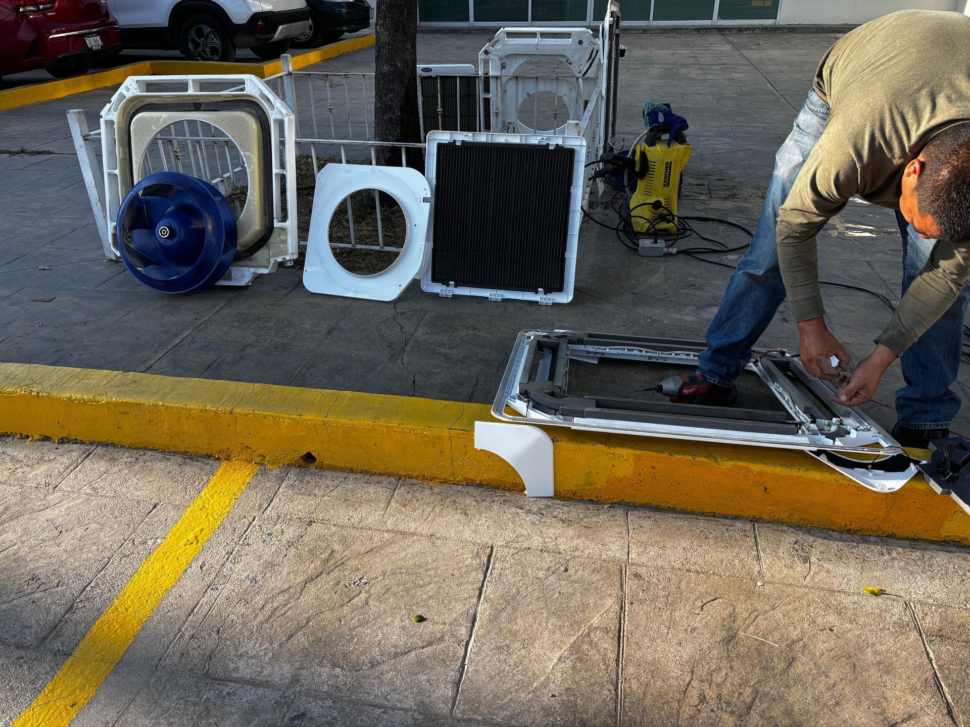 Un técnico desmonta una unidad de aire acondicionado en el bordillo de un aparcamiento, con las piezas esparcidas cerca.