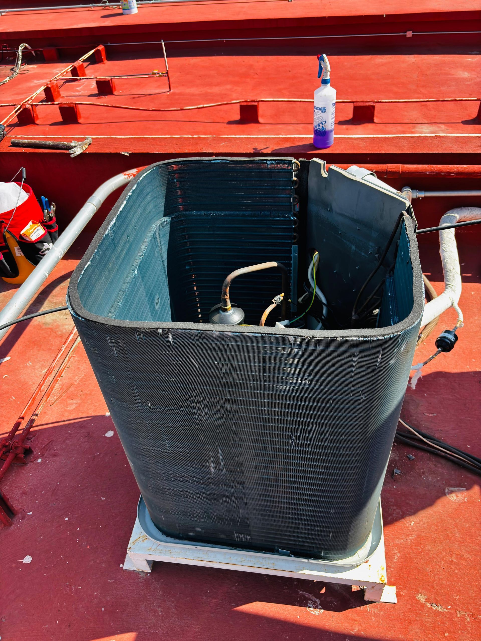 Una serpentina rectangular de condensador de climatización de color oscuro descansa sobre una base blanca en un techo rojo, con una botella de spray al fondo.